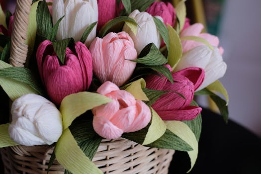 Panier en osier rempli de fleurs en papier crépon aux couleurs variées, roses et blanches, avec des feuilles vertes.